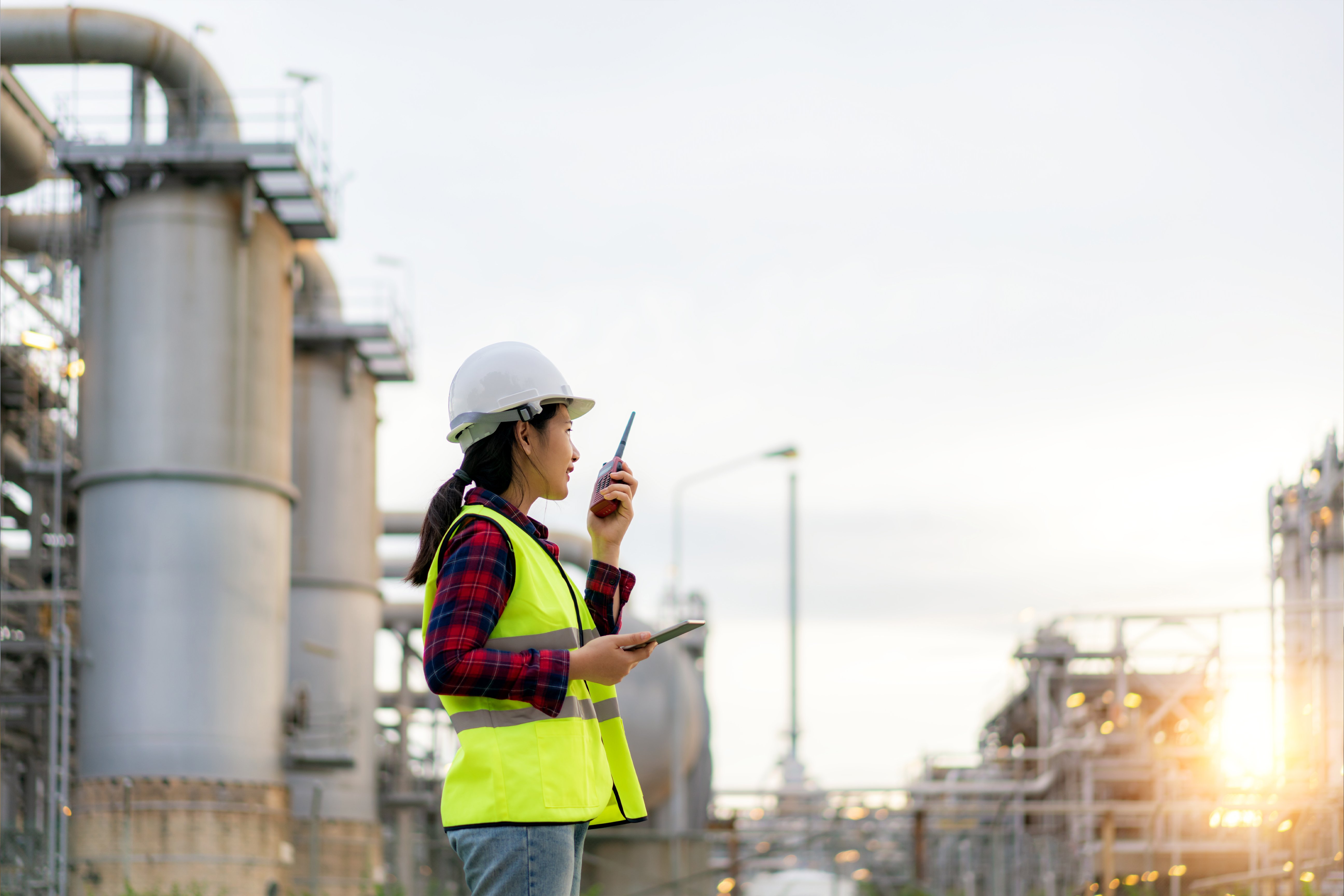 Asian woman technician Industrial engineer using walkie-talkie and holding bluprint working in oil refinery for building site survey in civil engineering project