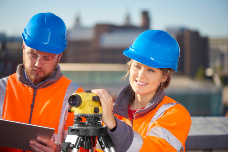 A female trainee surveyor stands at a builder's level supervised by her colleague, they are both smiling 